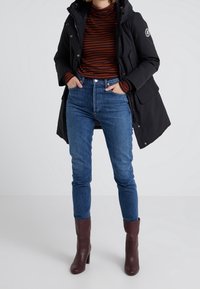 Sort parka med hætte, struktureret stof, orange og brune stribede turtleneck, blå tætsiddende denimbukser og brune hælede ankelstøvler.