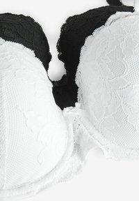 Soutiens-gorge rembourrés noirs et blancs avec un motif en dentelle florale, des bords festonnés et un petit nœud au centre où les bonnets se rejoignent.