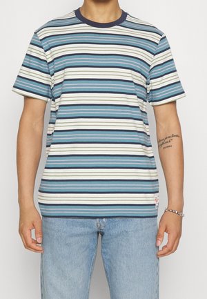 Mand iført en blå, hvid, sort og olivengrøn stribet T-shirt med korte ærmer, lys blå jeans og et sølvarmbånd på højre håndled.