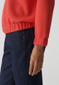 Rode textuurtrui met een geribbelde zoom en manchetten, gecombineerd met donkere denim jeans. De trui heeft een honingraatpatroon voor extra detail.