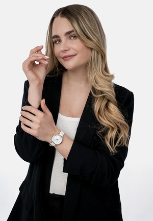 Junge Frau mit langen blonden Haaren trägt einen schwarzen Blazer, ein weißes Oberteil und eine weiße Armbanduhr, hält sanft ihr linkes Handgelenk mit der rechten Hand.