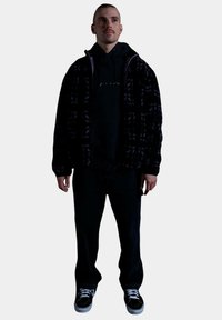 Chaqueta de plumas negra con una textura estampada, superpuesta sobre una sudadera negra con capucha. Unos jeans oscuros y zapatos negros completan el conjunto.