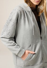 Sudadera con cremallera gris, confeccionada en una tela suave, con adornos de strass en los hombros y una capucha con cordón, además de bolsillos frontales.