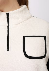 Φλις πουλόβερ σε κρεμ χρώμα με ψηλό γιακά, κλείσιμο με φερμουάρ και μαύρο διακοσμητικό στα τσέπες· διαθέτει απαλό υλικό και λιτό σχεδιασμό.