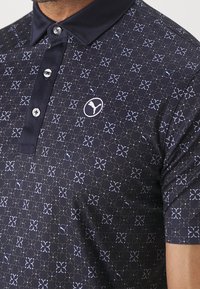 Marinblå polo-skjorta i mjukt tyg, med ett geometriskt mönster, knappad krage och en liten logotyp på bröstet.