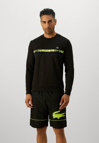 Svart hettegenser med en grønn og blå grafisk stripe. Kombinert med svarte shorts med et neongrønt krokodillelogo og detaljer.