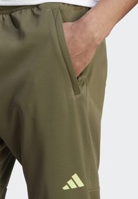 Olivenfarvede træningsbukser med en struktureret overflade, som har en sidelomme og et reflekterende gult Adidas-logo på underbenet.