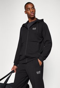Black zip-up hoodie with hood, featuring a logo on the chest, paired with matching black joggers. Smooth fabric, minimalistic design.