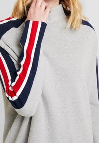 Jasnoszary sweter z golfem o prążkowanych granatowych rękawach z czerwonymi, białymi i granatowymi paskami. Miękka tekstura i luźny krój.