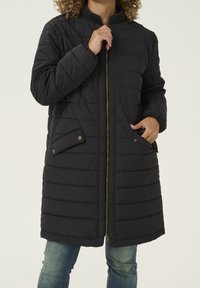 Abrigo negro acolchado con cierre de cremallera en el frente, cuello acanalado y dos bolsillos laterales con cierres de botón a presión. Textura suave y diseño hasta la rodilla.