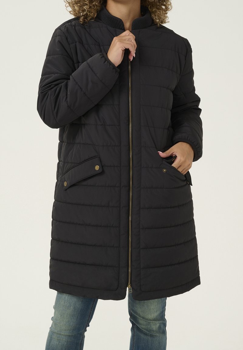 Abrigo negro acolchado con cierre de cremallera en el frente, cuello acanalado y dos bolsillos laterales con cierres de botón a presión. Textura suave y diseño hasta la rodilla.