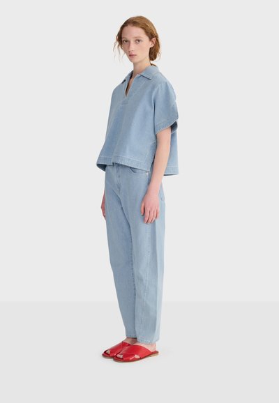 Jeune femme portant un ensemble ample en denim bleu clair, debout de profil, avec des sandales rouges plates à bout ouvert, sur un fond clair uni.