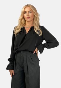 Czarna satynowa bluzka z dekoltem w serek i marszczonymi mankietami, zestawiona z wysokim stanem ciemnych spodni w szerokie paski. Złoty łańcuszek akcentujący dekolt.