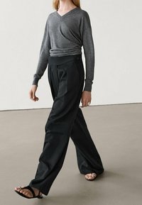 Grijze v-hals trui met lange mouwen, gecombineerd met zwarte wijde broek. De outfit heeft een ontspannen pasvorm en een zachte textuur.