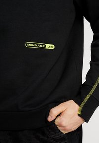 Sudadera negra con una textura suave, que presenta un logo verde "MENNACE LTD" en el frente y un acento de costura amarilla en la manga.