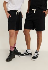 Černé šortky vyrobené z bavlněné směsi, s elastickým pasem a šňůrkou na stažení. Dva jednotlivci nosí různé obuv: černé boty a tenisky.