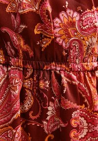 Tissu marron avec un motif cachemire en orange, rose et blanc. Présente des détails froncés à la taille avec une texture lisse.