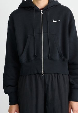 Personne portant un sweat à capuche zippé noir Nike avec cordons de serrage et poches kangourou, assorti à un pantalon noir ample.