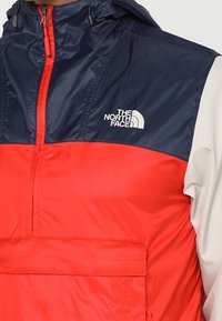 Veste coupe-vent rouge et marine avec capuche, fabriquée en matériau léger. Comprend une poche avant et un logo de marque sur la poitrine.