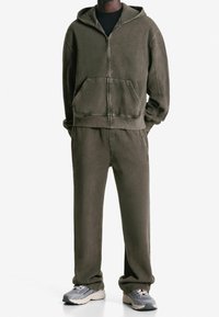 Conjunto a juego de sudadera con capucha y pantalones deportivos en verde oliva, confeccionado con tela suave. La sudadera con capucha tiene cremallera frontal y bolsillo tipo canguro. Los zapatos son zapatillas grises.