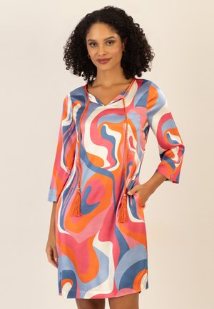 Frau mit lockigem Haar, die ein buntes Kleid mit abstraktem Wirbelmuster, 3/4-Ärmeln und orangen Quastenbändern am Ausschnitt trägt.