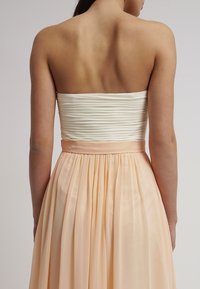 Femme portant une robe bustier avec un corsage blanc froncé et une jupe plissée couleur pêche douce, vue de dos sur fond neutre.