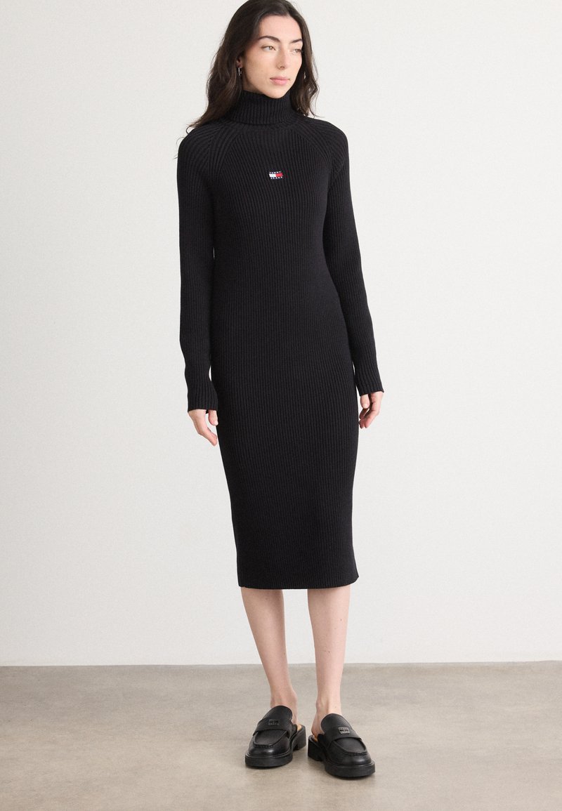 Musta ribbed turtleneck-mekko, jossa on pitkät hihat ja polvipituinen muotoilu. Mekossa on pieni logo rinnassa ja se istuu tiiviisti.