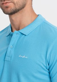 Vaaleansininen poolopaita, jossa on tekstuuri, tavanomainen kaulus, kaksi nappia ja pieni valkoinen Brodeerattu logo rinnassa.