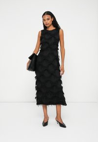 Mujer vestida con un vestido midi negro sin mangas con patrones circulares texturizados, sosteniendo un bolso de mano negro y usando mules de tacón negros.