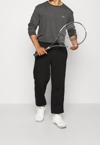 Szara koszula z długim rękawem z subtelnym logo, czarne spodnie i białe buty sportowe. Trzyma rakietę z metalową ramą i naciągiem z siateczki.