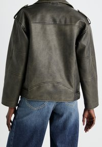 Zielona kurtka z ekologicznej skóry z ostrym kołnierzem, pagonami na ramionach i subtelnymi detalami przeszyć, zestawiona z szerokimi niebieskimi dżinsami.