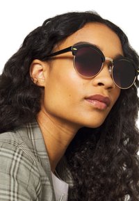 Gafas de sol redondas con montura negra y lentes oscuros con degradado. Presentan acentos dorados y un diseño único en la parte superior de los lentes.