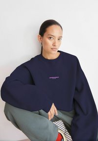 Marineblaues Sweatshirt mit übergroßen Ärmeln und Rundhalsausschnitt, mit dem "ARMEDANGELS" Logo in Pink. Getragen zu hellgrünen Hosen und gestreiften Schuhen.