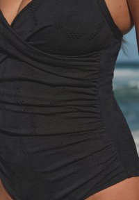 Must ujumistrikoo tekstuurse kangaga, millel on peen muster, külgedel kogutud disain ja reguleeritavad rihmad sobivuse tagamiseks.