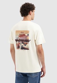 Crème-kleurig katoenen t-shirt met een grafische print op de achterkant, met abstracte kunst, tekst in rood en bruin, korte mouwen.
