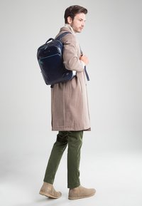 Mochila de cuero azul marino con acentos en azul claro, forma rectangular, correas ajustables y un acabado suave, llevada sobre un abrigo beige y pantalones verdes.