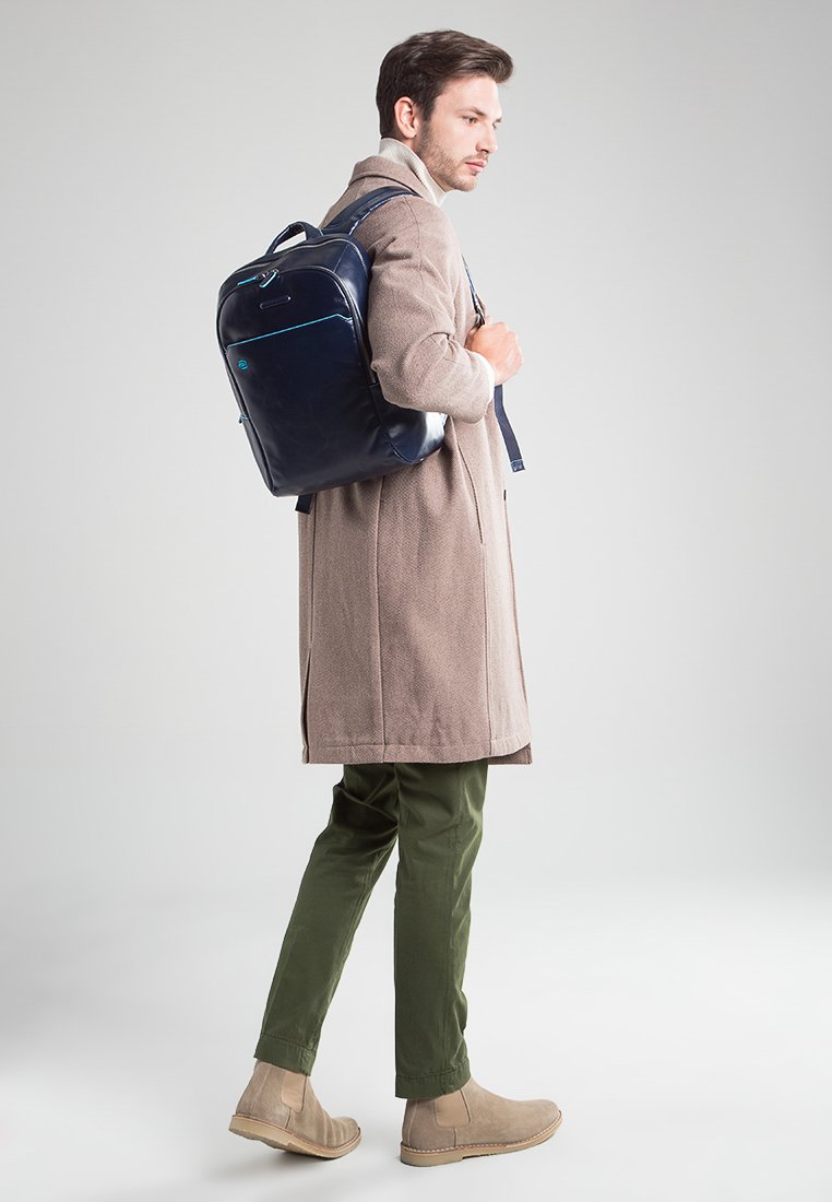 Mochila de cuero azul marino con acentos en azul claro, forma rectangular, correas ajustables y un acabado suave, llevada sobre un abrigo beige y pantalones verdes.