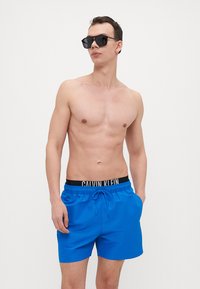 Blaue Badehosen aus glattem Stoff mit einem Kordelzugbund und einem schwarzen Logoband. Modell trägt schwarze Sonnenbrille und kein Shirt.