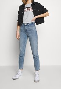 Žena v světle modrých vysoce pasových džínách, šedém tričku s grafikou, černé zkrácené bundě a bílých vysokých teniskách stojí proti bílému pozadí.