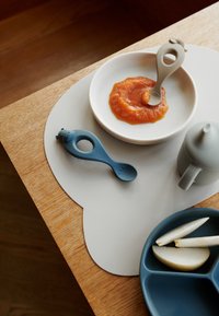 Blue silicone spoon with bear design, white bowl with orange puree, gray cup, and divided blue plate with pear slices on a soft gray placemat.