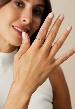 Guldbåndsring med et tekstureret, solstråleagtigt design på toppen, båret på en hånd med lys hud og velplejede, nude negle.