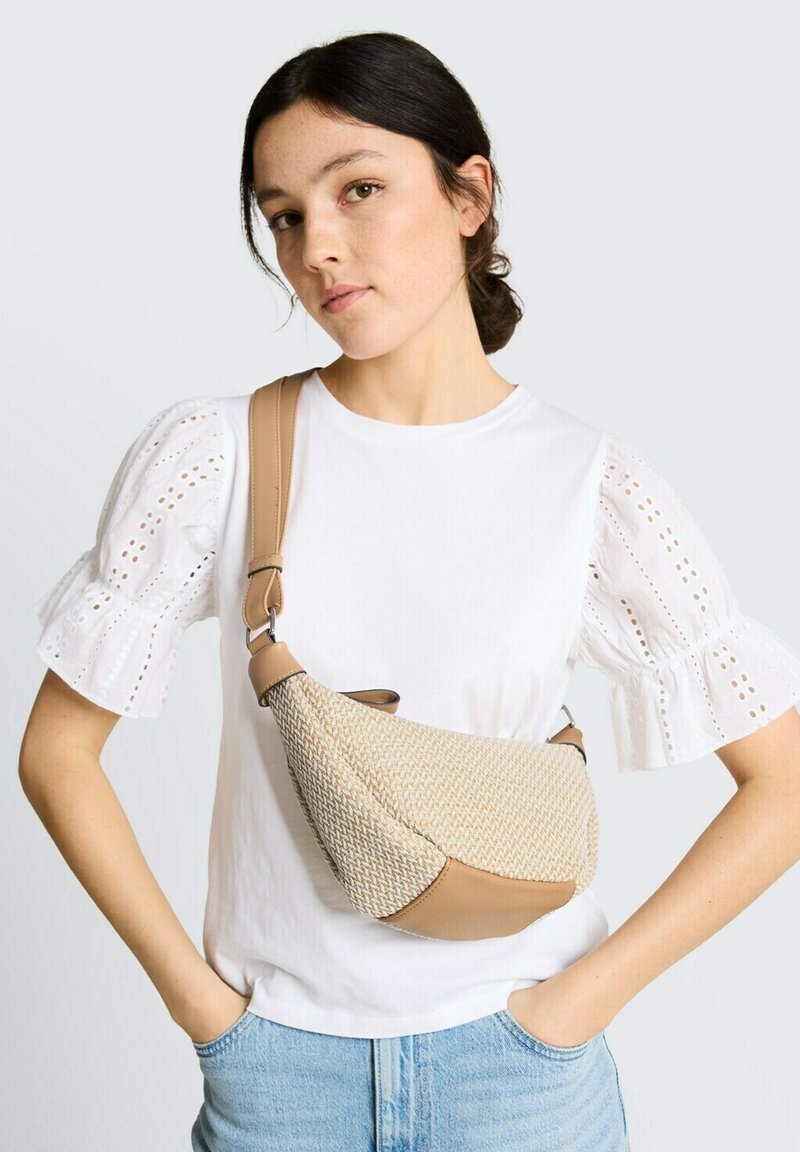 Jeune femme portant un chemisier blanc à manches courtes en œillet et un jean bleu, portant un sac bandoulière beige en tissu tressé avec des accents en cuir.