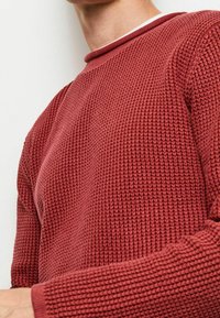 Roter Strickpullover mit strukturiertem Muster, Rundhalsausschnitt und gerippten Bündchen. Der Stoff wirkt warm und lässig.