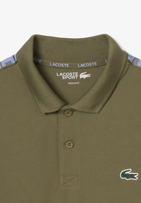 Polo verde oliva realizzato in tessuto strutturato, con tre bottoni, colletto e un piccolo logo ricamato a forma di coccodrillo sul petto. Presenta dettagli a fantasia.