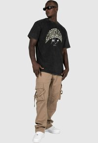 Svart grafisk t-skjorte med hvit skrift, kombinert med beige cargo-bukser med flere lommer og knytedetaljer; hvite joggesko.