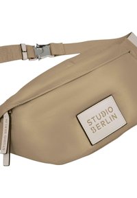 Béžový látkový pouzdro na pás s hladkou textúrou, s obdlžníkovou bielou nálepkou značky "STUDIO BERLIN" a nastaviteľným popruhom s kovovou sponou.
