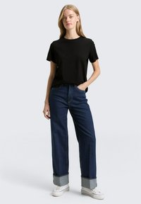 Schwarzes, kurzärmeliges T-Shirt mit erhabenem Text, kombiniert mit hochgezogenen, weit geschnittenen Dunkelblauen Jeans mit aufgerollten Säumen und weißen Sneakern.