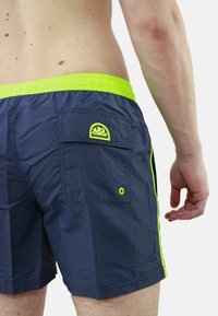 Pantaloni da bagno blu navy con passante elastico verde brillante. Le caratteristiche includono una tasca sul retro con un logo coordinato e strisce laterali a contrasto.