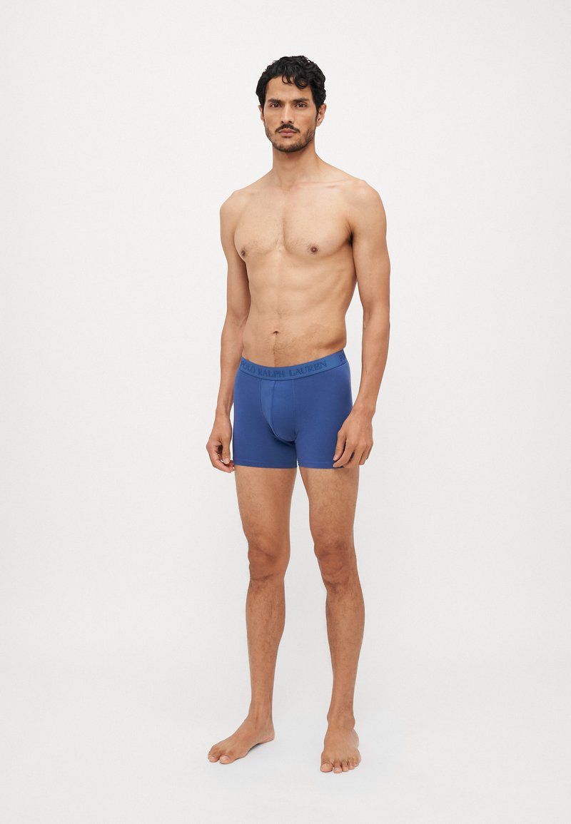 Blaue Stretch-Baumwollboxershorts mit elastischem Bund, eng anliegendem Design und Oberschenkellänge, auf einem nackten männlichen Modell vor einem weißen Hintergrund.