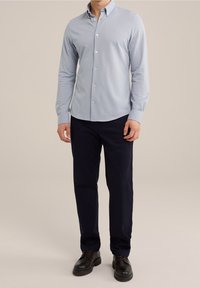 Světlomodrá košile na knoflíky s dlouhými rukávy, v kombinaci s tmavě modrými kalhotami a černými botami. Košile má jemný strukturovaný vzor.